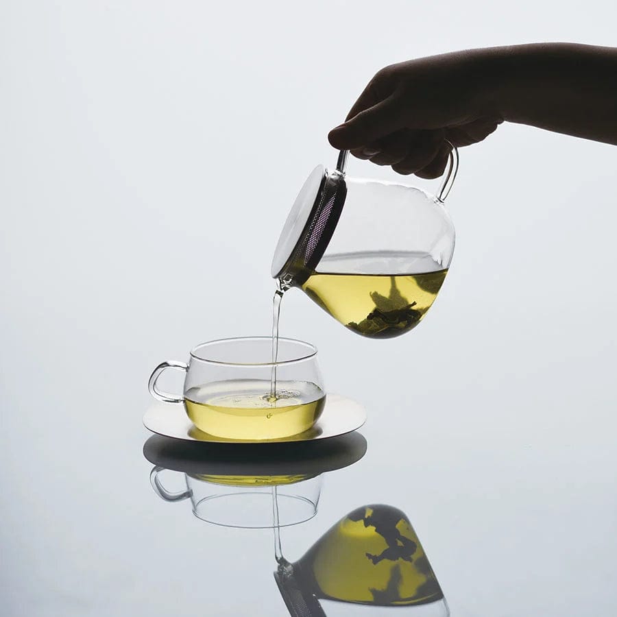 A hand pours green tea from a glass teapot into a matching cup and saucer, with a reflection below.