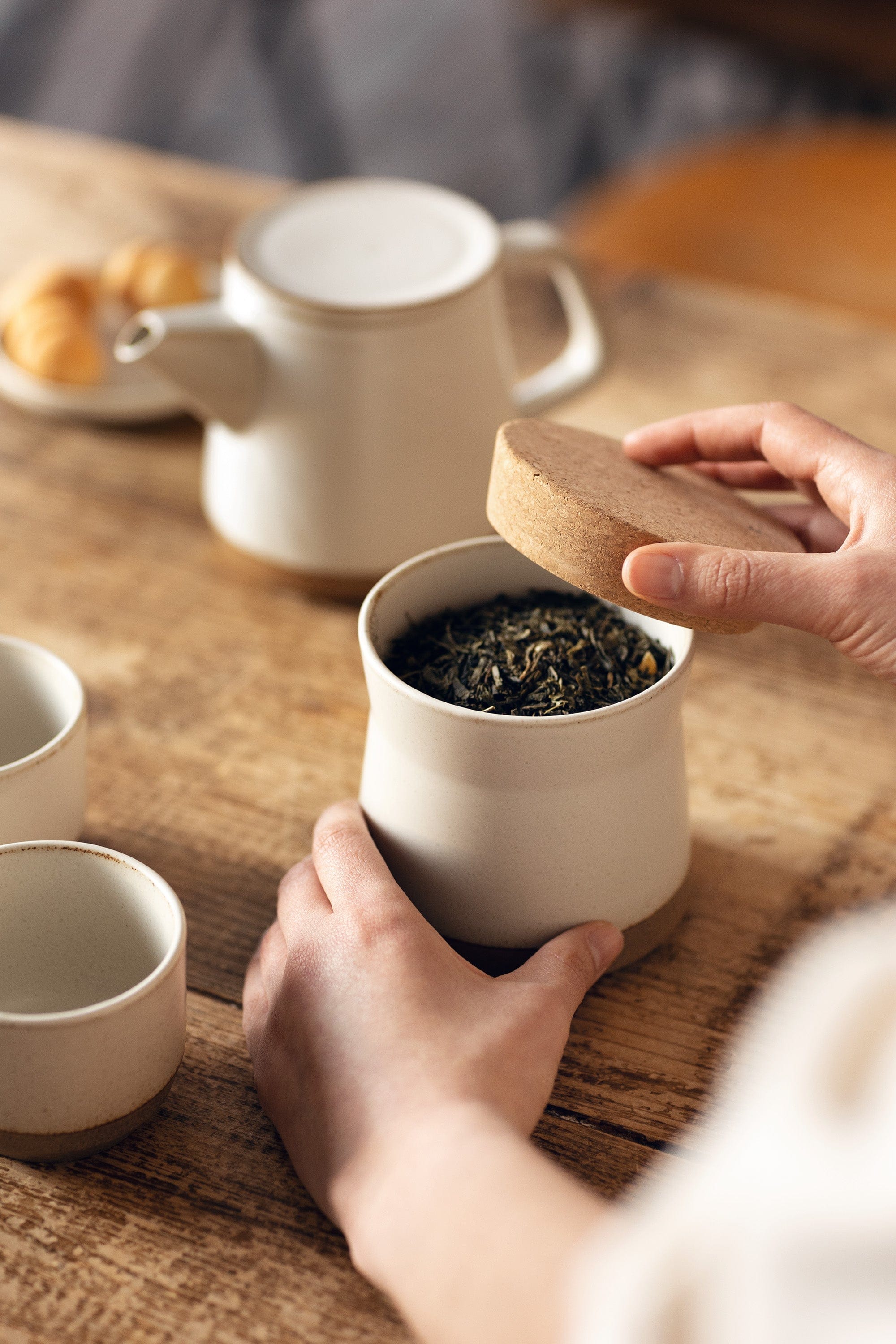 Person opening a ceramic jar filled with loose tea, on a wooden table with cups and a teapot.