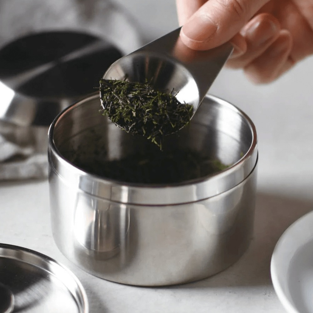 Hand scooping dry tea leaves from a metal canister with a spoon.