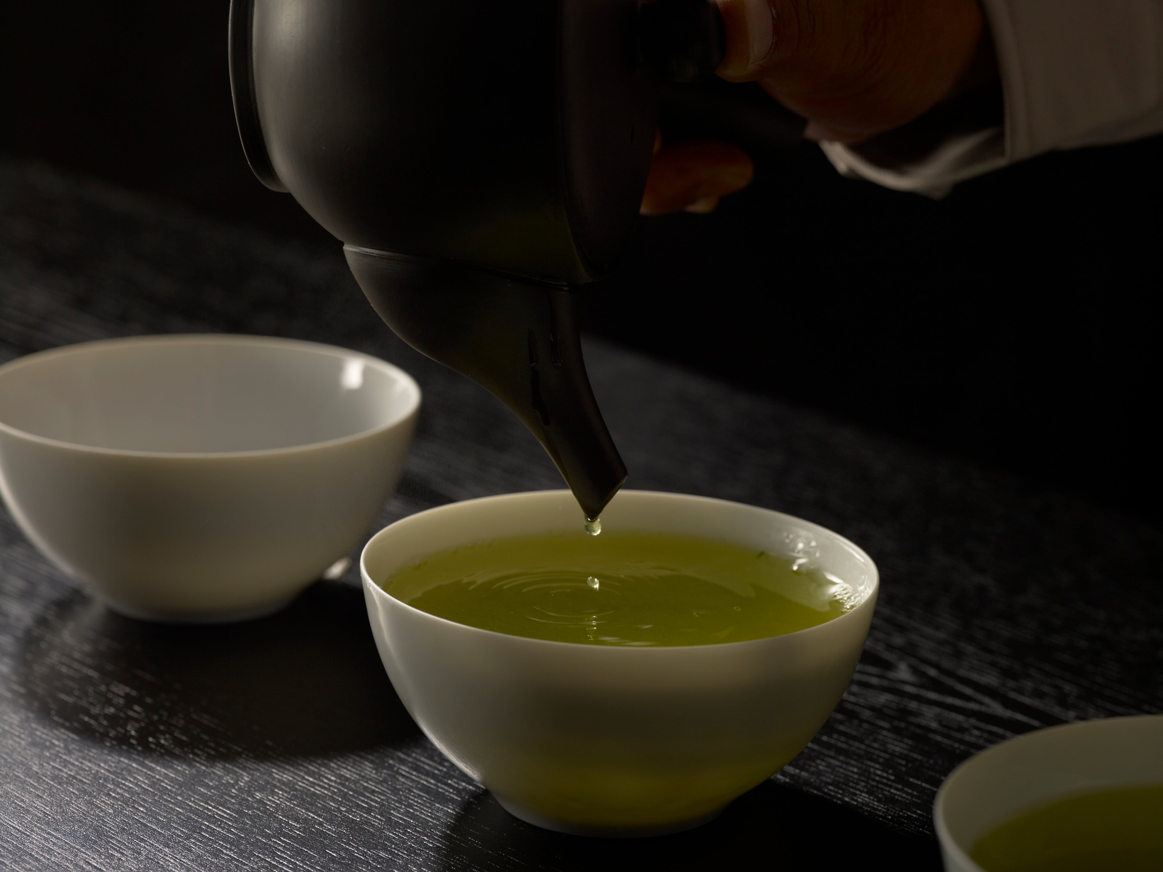 Pouring green tea from a teapot into a white bowl on a dark wooden table.