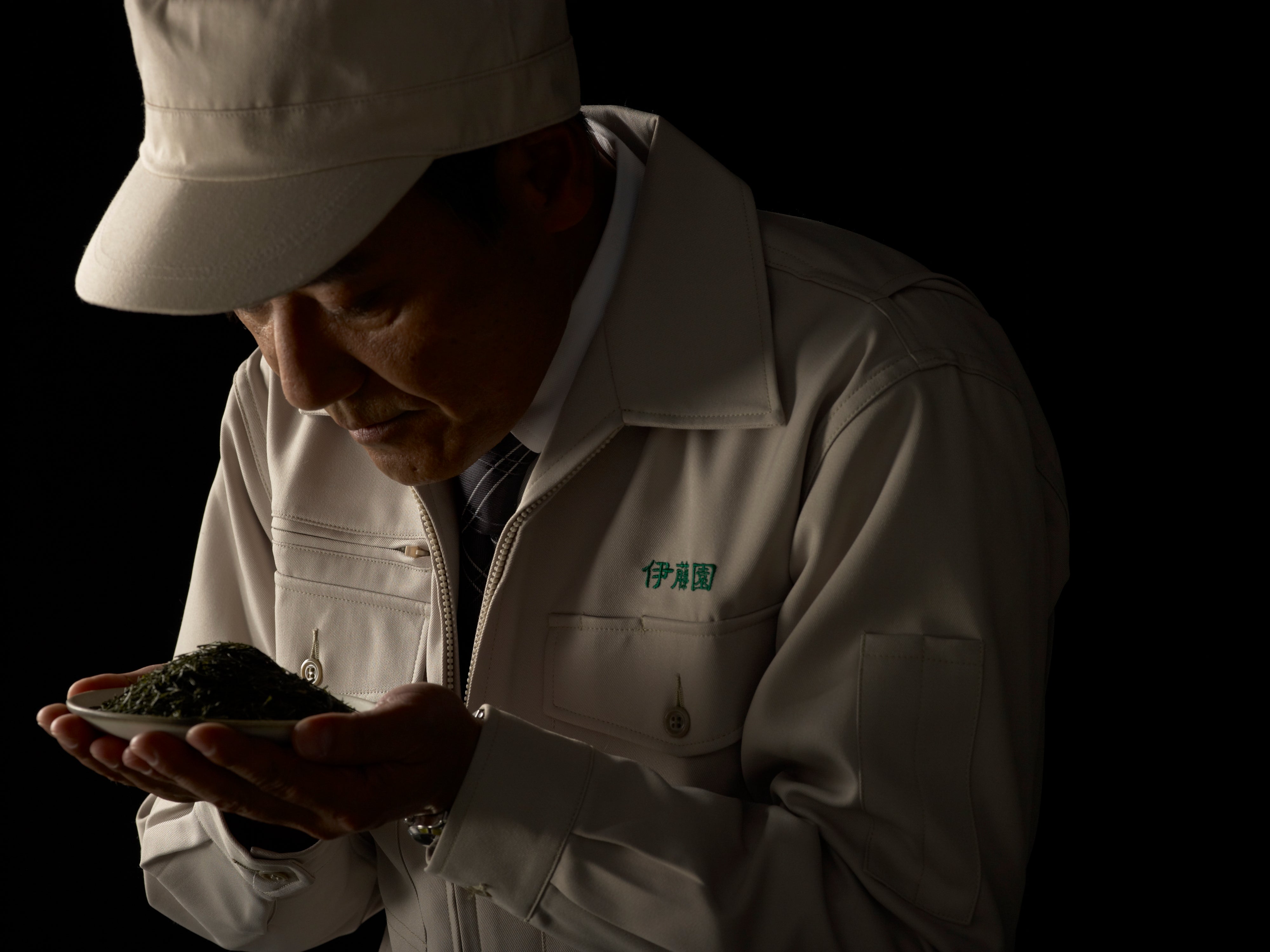Person in a beige jacket and cap smelling green tea leaves in their hands.