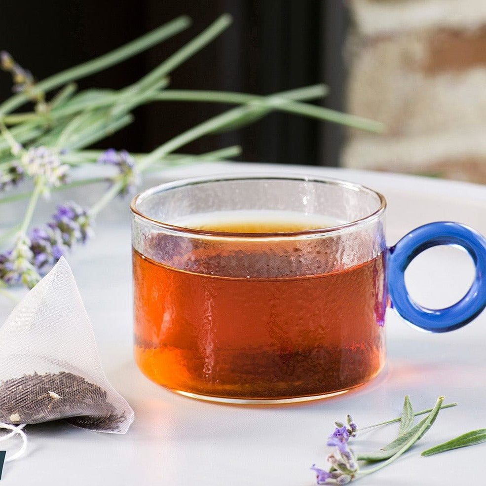 Glass cup of dark tea with a blue handle beside a tea bag and lavender sprigs.