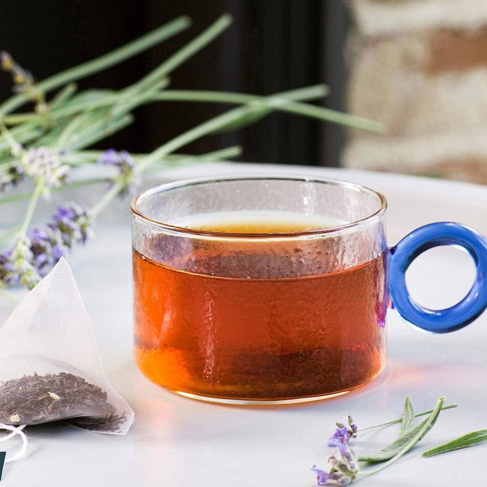 Glass cup of tea beside a tea bag and sprigs on a white surface.