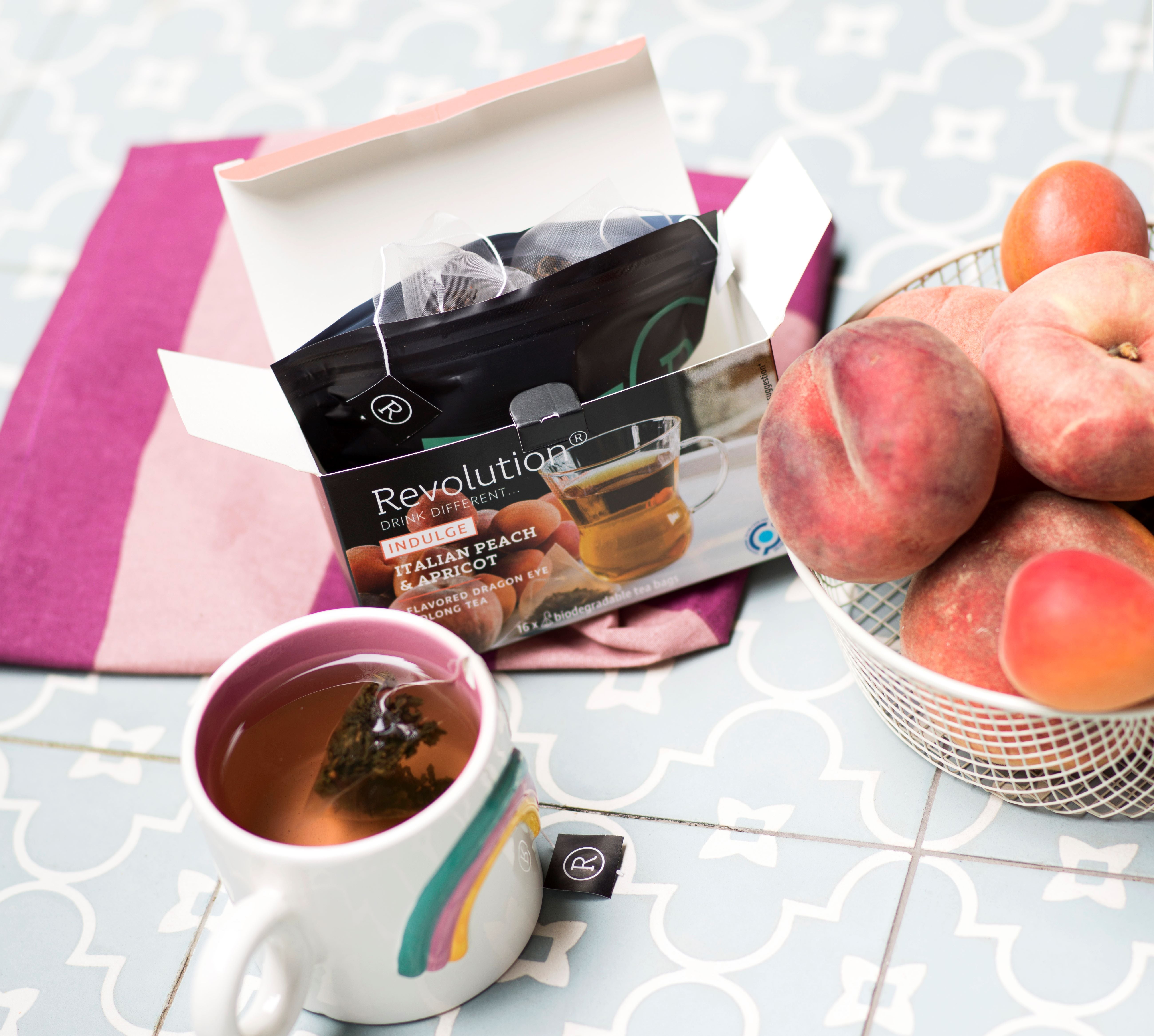 Tea box, rainbow mug, and fresh peaches on a blue tile surface.