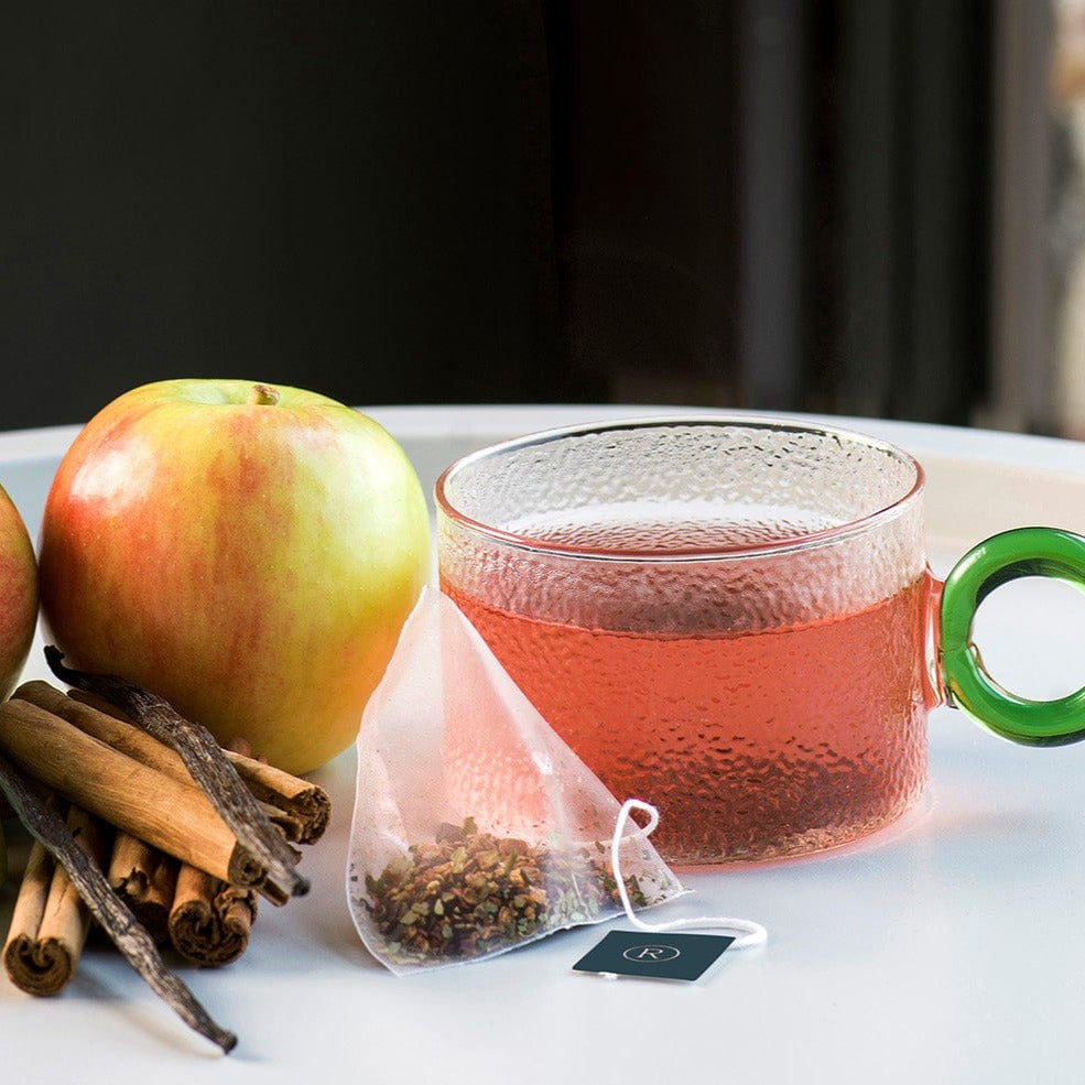Clear mug of red tea, apple, cinnamon sticks, and tea bag on a white surface.
