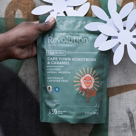 Hand holds Cape Town Honeybush & Caramel herbal infusion pouch; paper flowers.