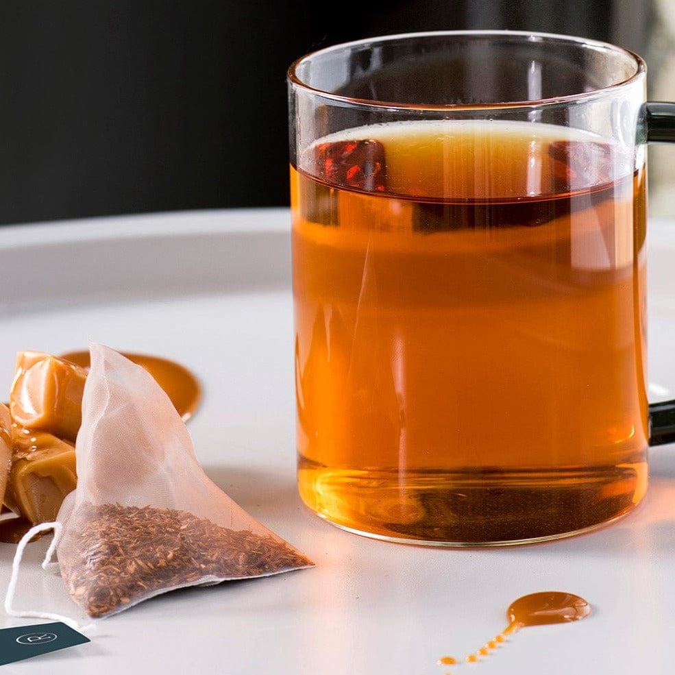 Glass mug with brown tea beside caramel and a tea bag on a white surface.