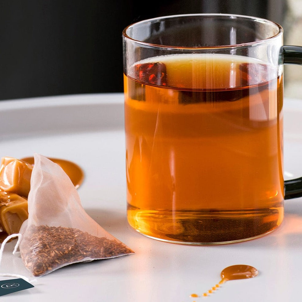 Clear mug of amber tea, ginger slices, cookies, and tea bag on a white surface.
