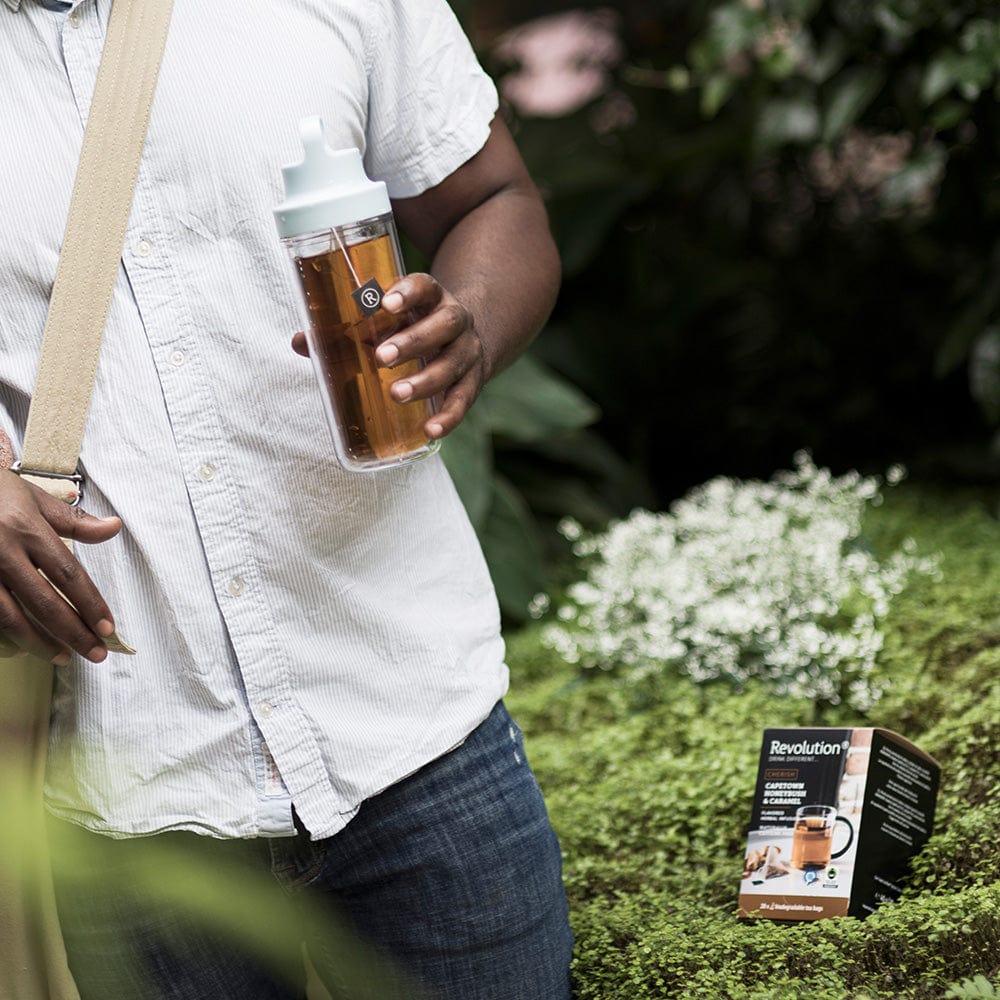 Person in white shirt holding tea bottle with a pouch outside in a garden.