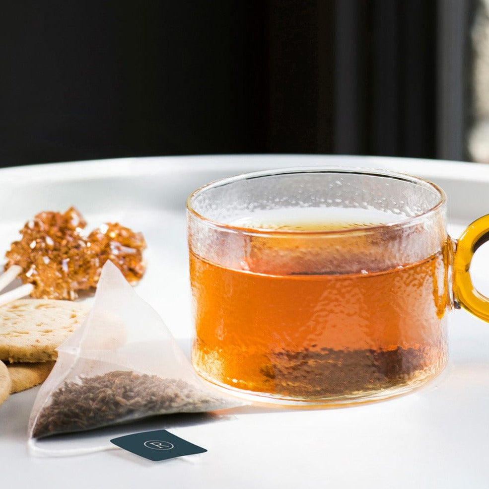 Clear glass mug of amber tea with ginger slices, cookies, and a tea bag on a tray.