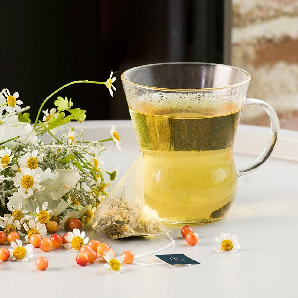 Clear mug of yellow tea with chamomile flowers, orange berries, and a tea bag nearby.