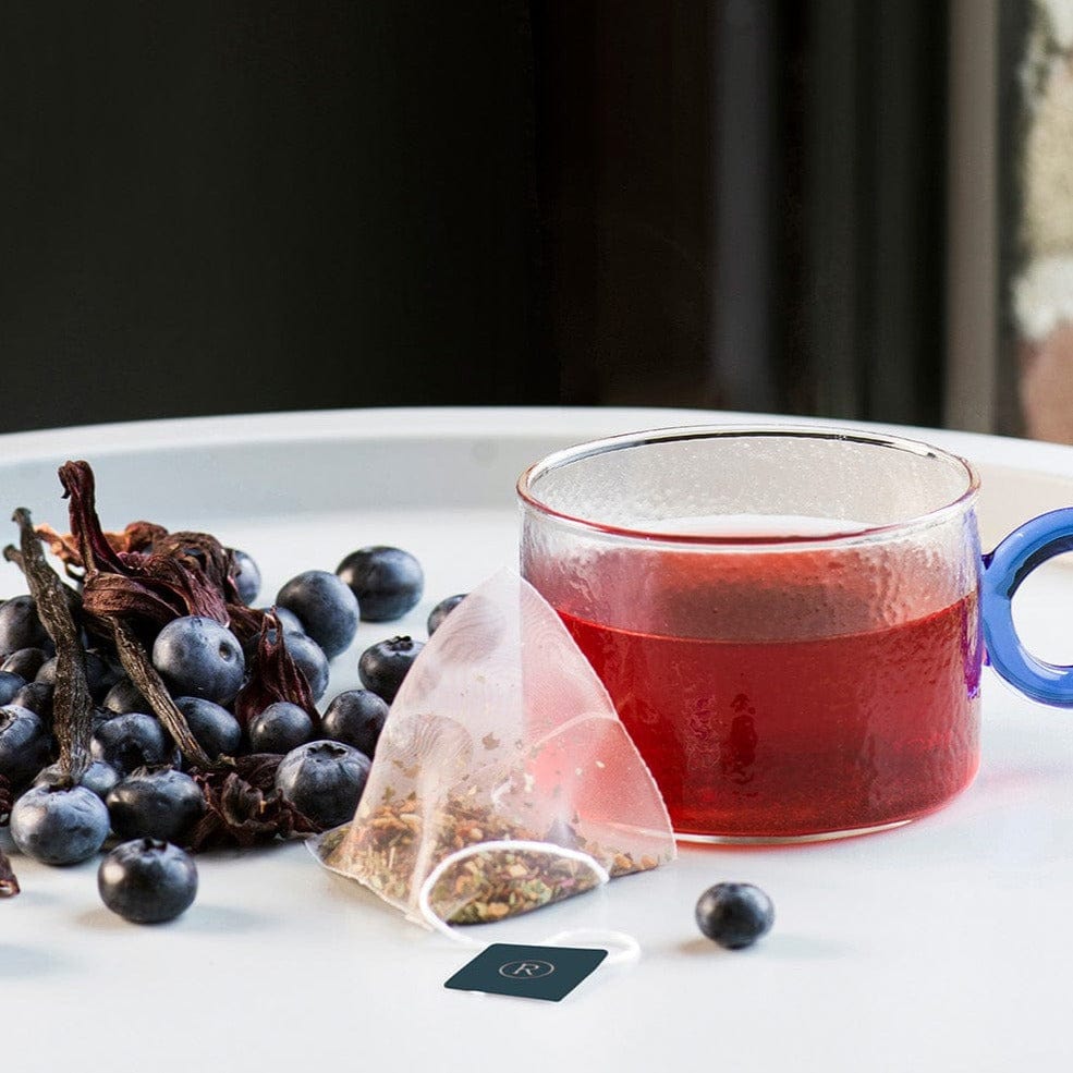 Glass mug with red tea beside blueberries and tea bag on a white tray.