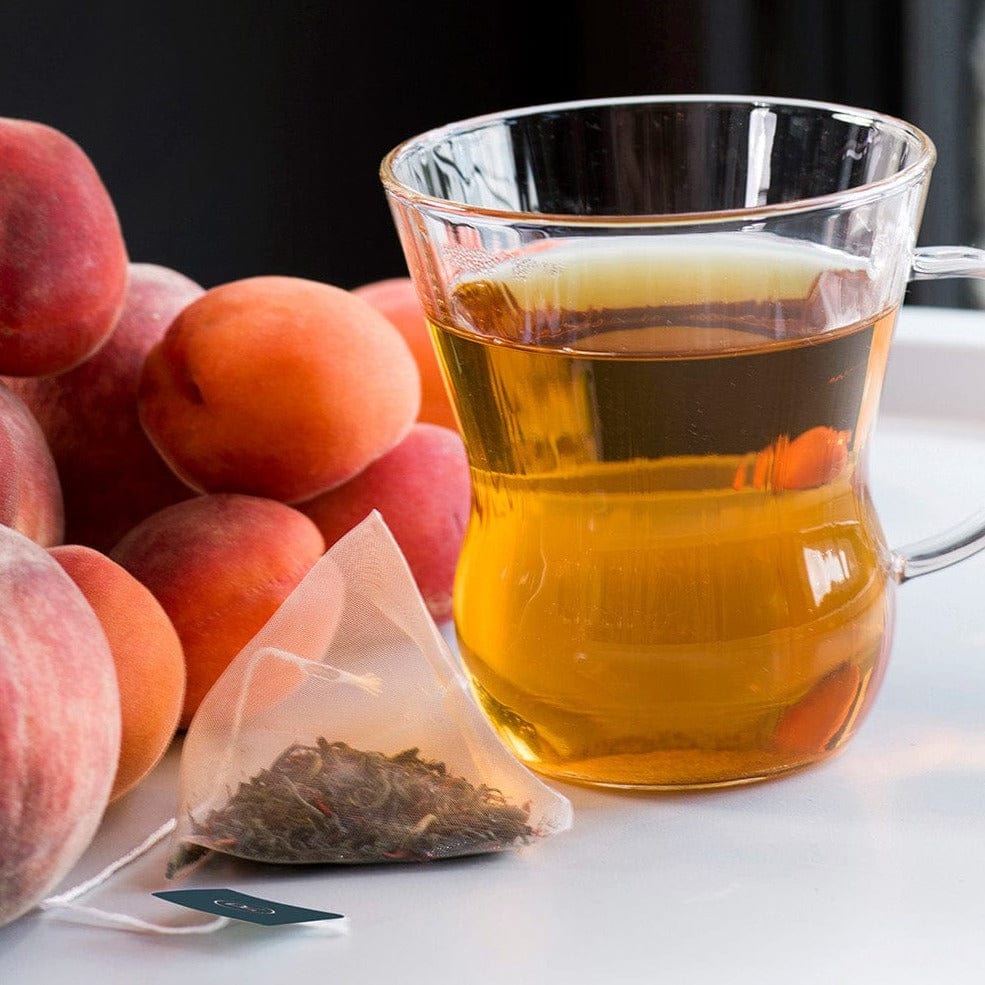 Glass of amber tea next to whole peaches and a tea bag on a white tray.