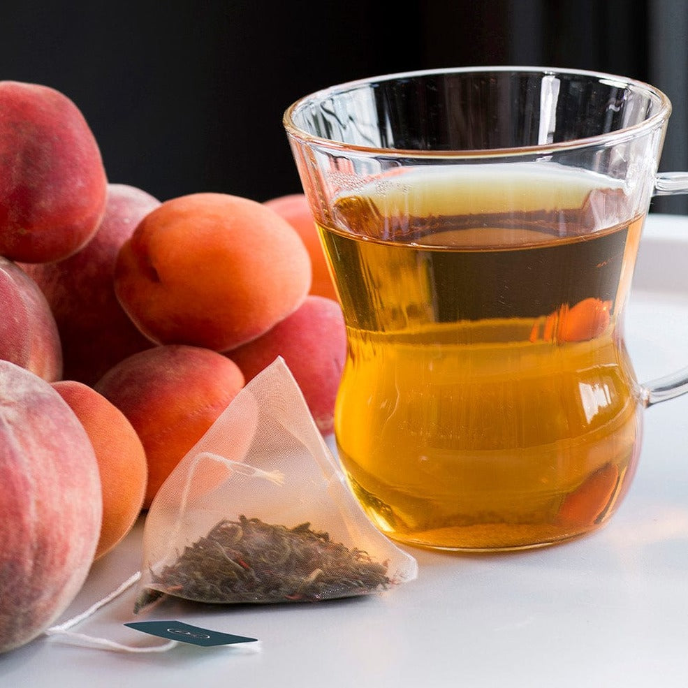 Clear mug with golden peach tea and a pyramid tea bag, peaches beside it.