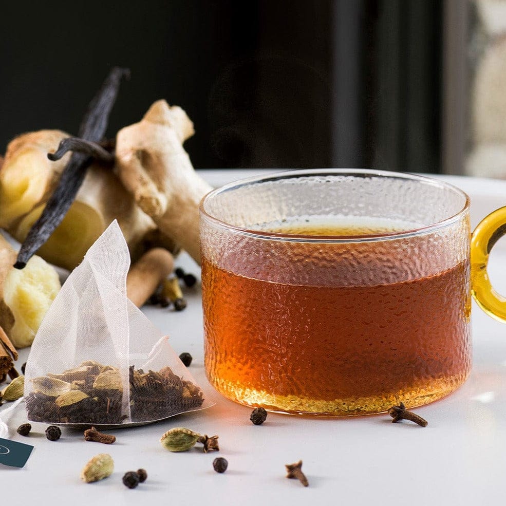 Glass cup of tea with cardamom, ginger, vanilla, and a tea bag on the side.