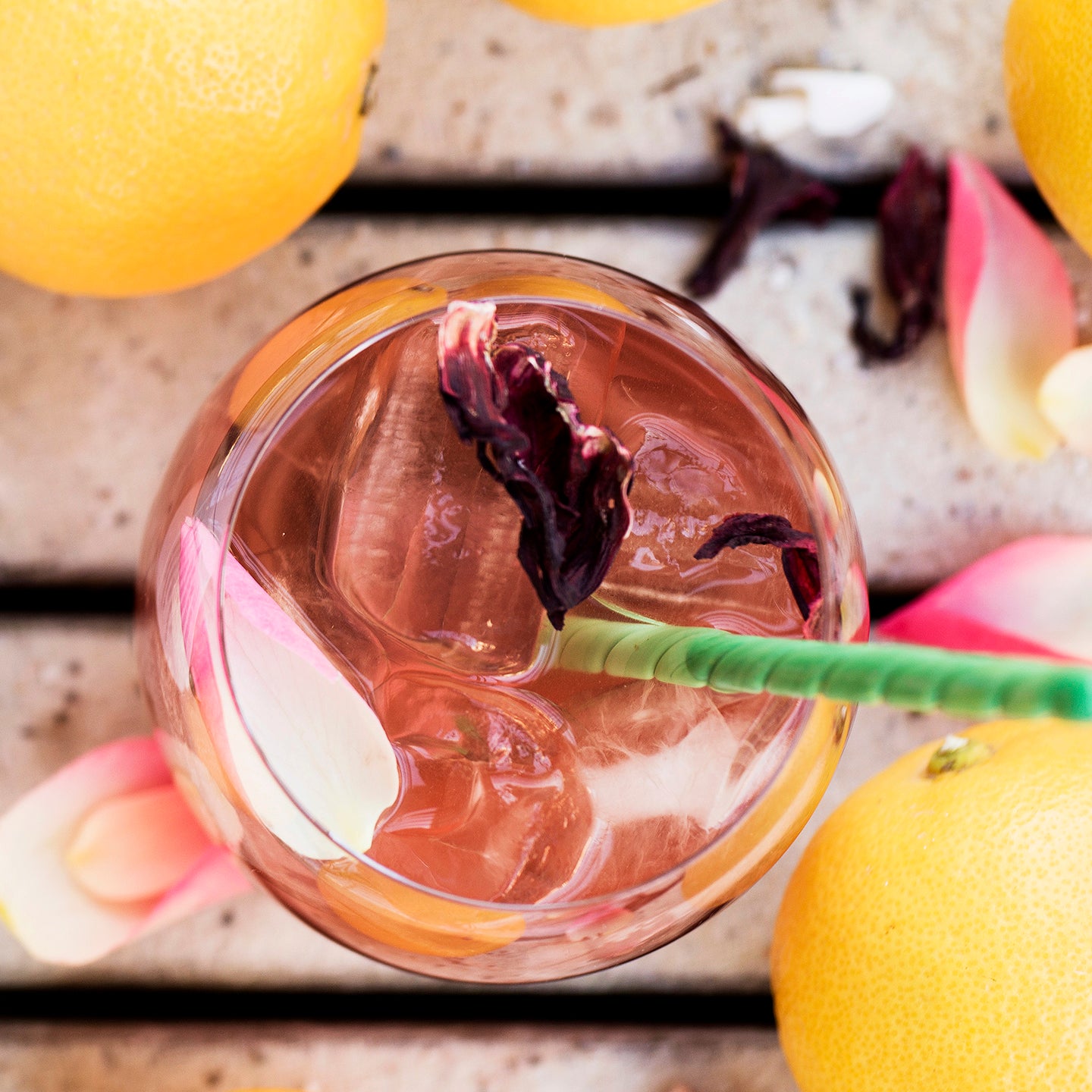 Pink iced tea with a straw in a glass surrounded by lemons and rose petals.