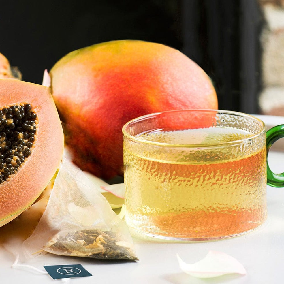 Glass mug with light amber tea, papaya and mango halves, and a tea bag on a white table.