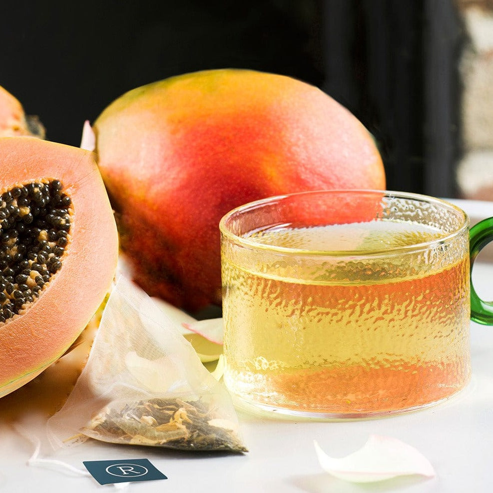 Glass mug with light amber tea, papaya and mango halves, and a tea bag on a white table.