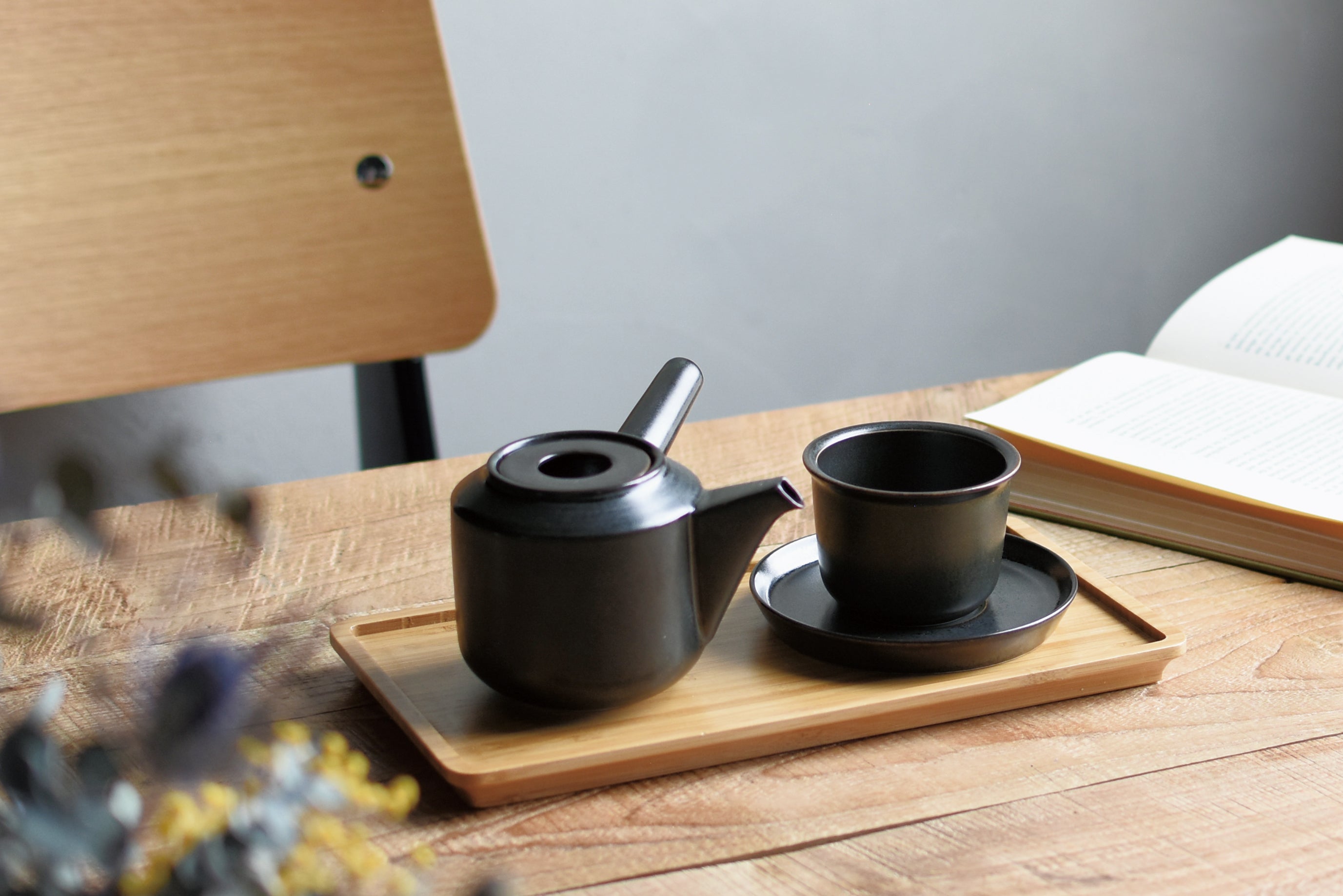 Black teapot and cup on a wooden tray with an open book in the background.