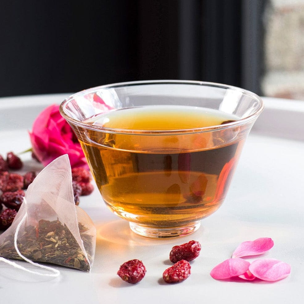 Cup of amber tea with cranberries, rose petals, and a tea bag on a white tray.