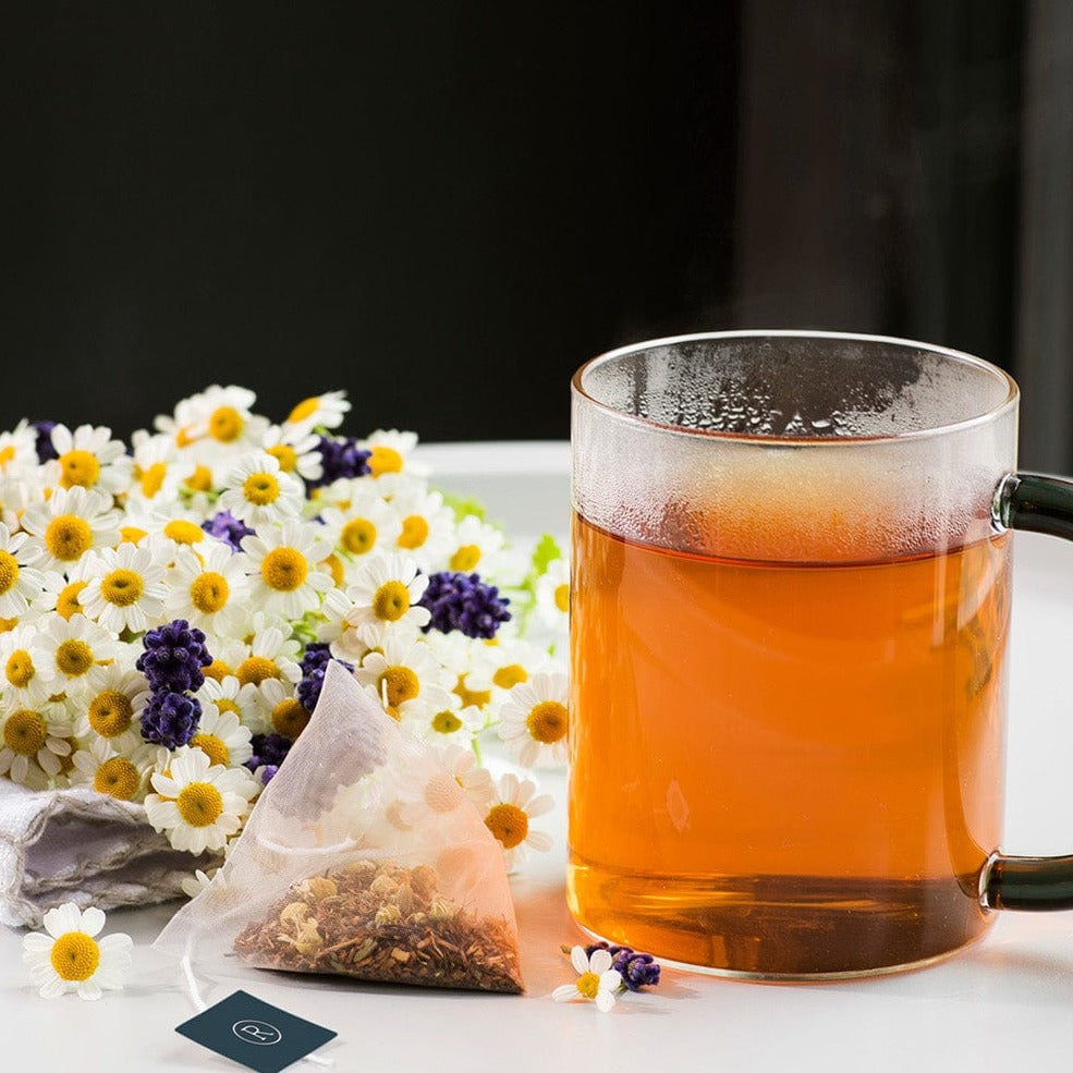 Glass mug with dark amber tea surrounded by white and yellow flowers and a tea bag.