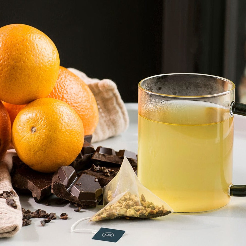 Glass mug with pale tea, surrounded by oranges, chocolate, and a tea bag on white table.