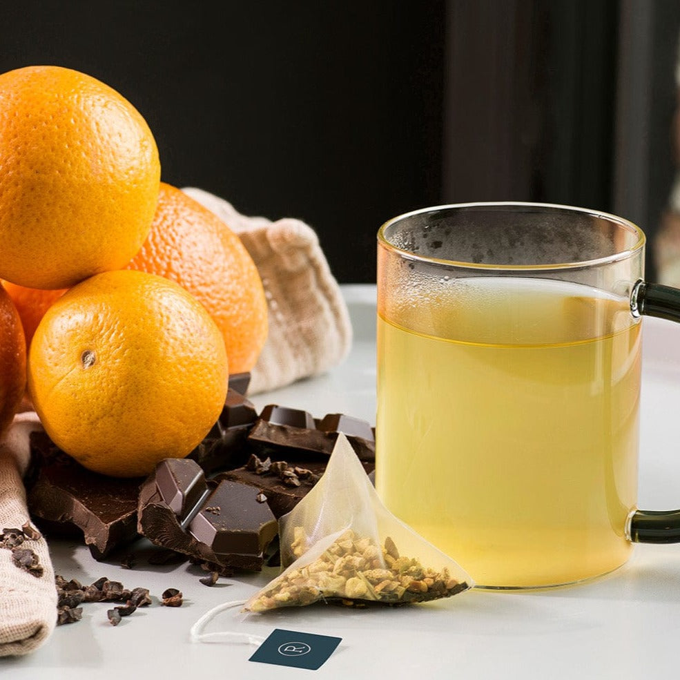 Glass mug with pale tea, surrounded by oranges, chocolate, and a tea bag on white table.