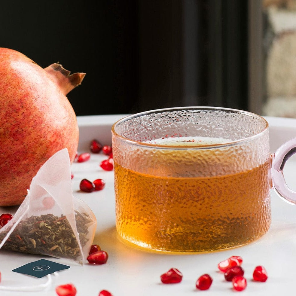 Glass mug with amber tea surrounded by a whole pomegranate, seeds, and a tea bag.
