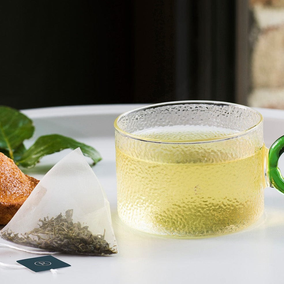Clear glass mug with light green tea beside a triangular tea bag on a white tray.