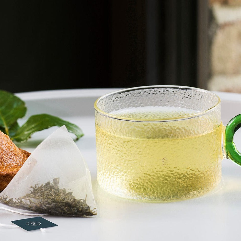 Clear glass mug with amber tea next to a triangular tea bag and mint leaves.