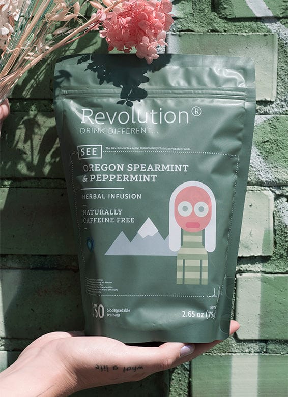 Hand holds Oregon Spearmint & Peppermint herbal infusion pouch; pink flowers above.