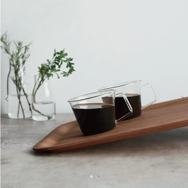Two glass cups with dark liquid on a wooden tray, plants in glass vases in the background.