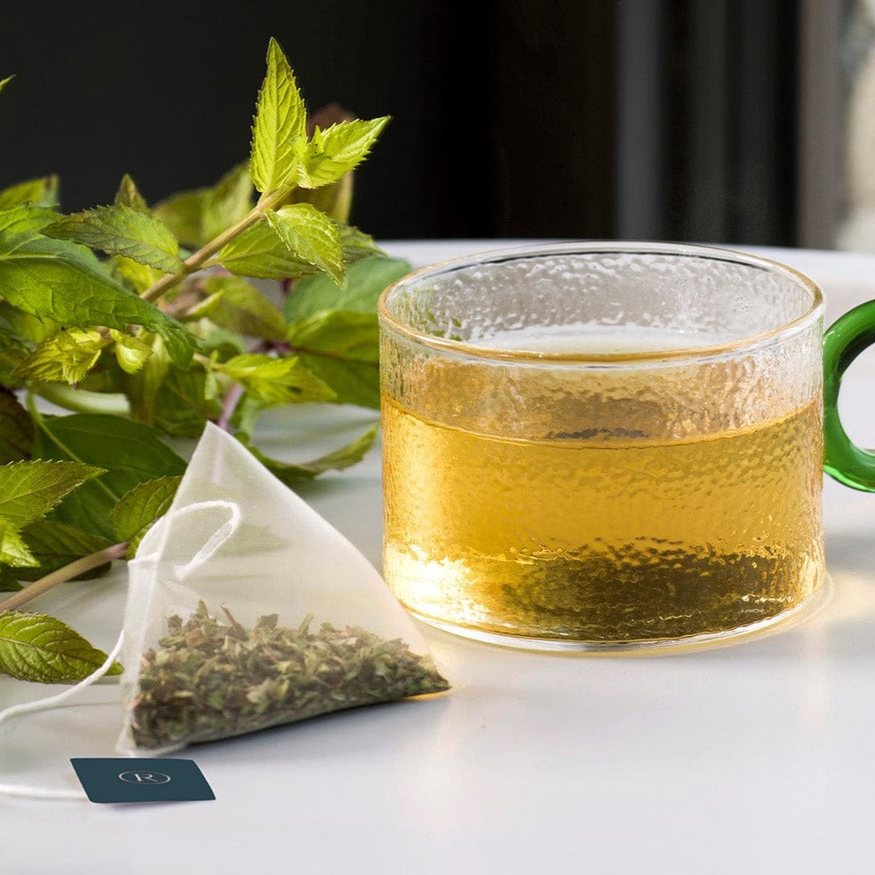 Glass mug with peach tea next to peaches and a triangular tea bag on a tray.