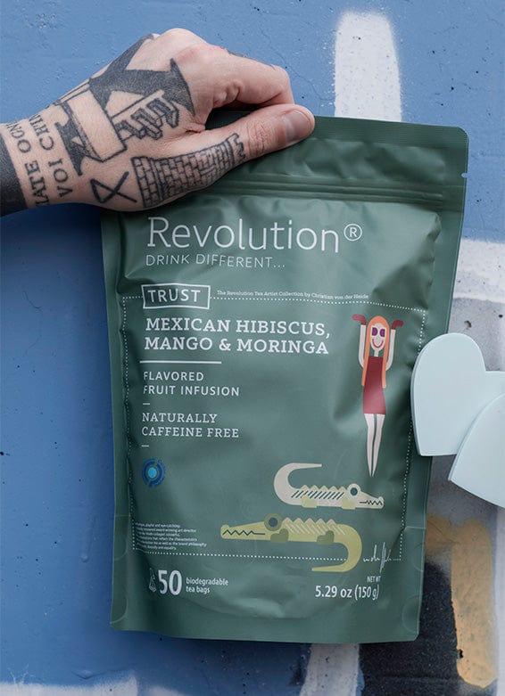 A tattooed hand holds a green tea pouch labeled 'Revolution' with hibiscus, mango, and moringa.