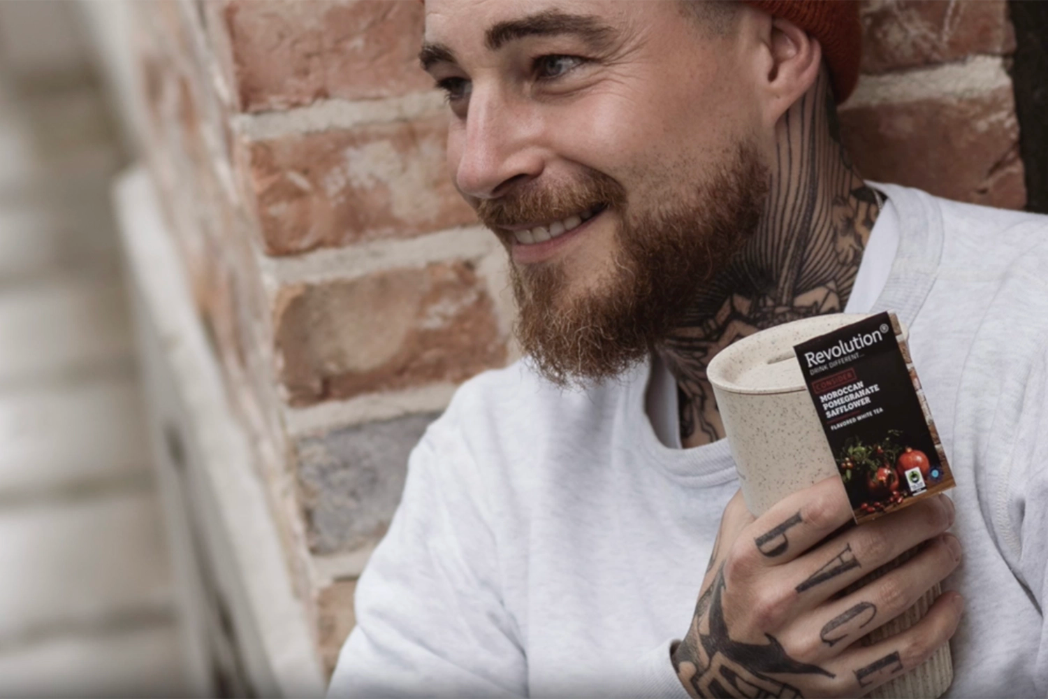 Man with tattoos holds a beige mug, smiling against a brick wall.