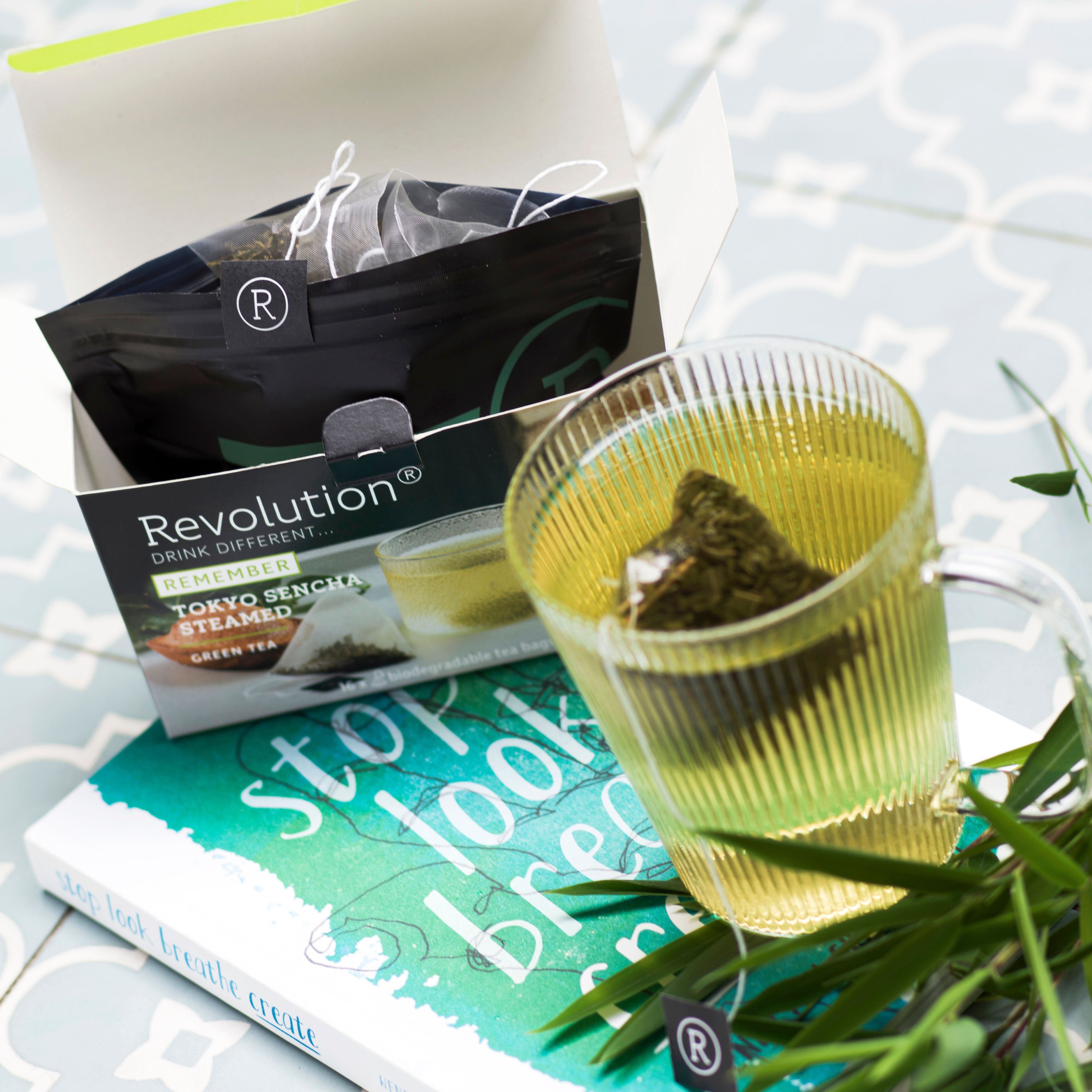 Glass of green tea with bamboo, a book, and a tea box on a tiled table.