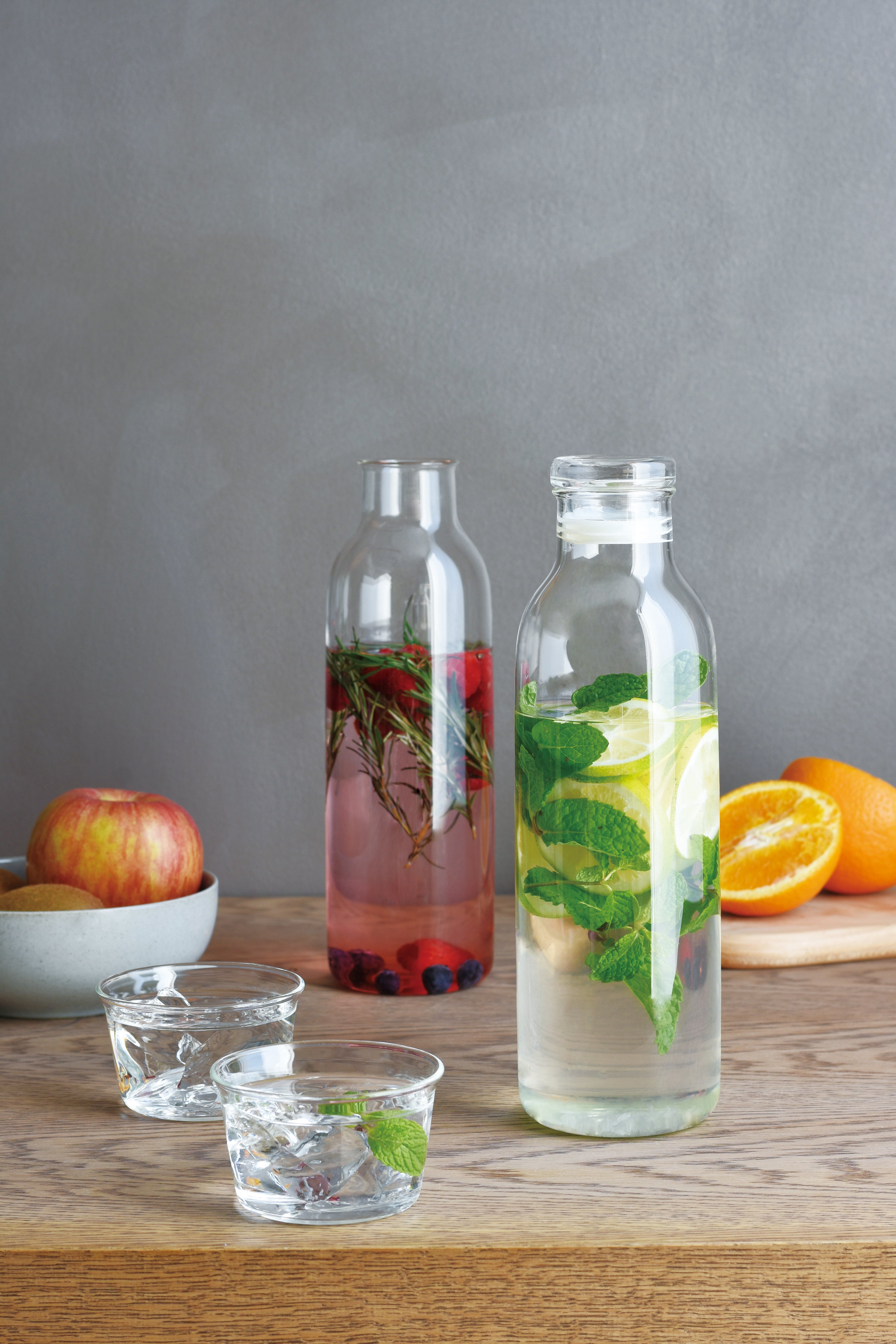 Two glass bottles on a wooden table with fruits, both filled with infused water.