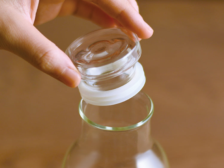 Hand placing a transparent lid on a glass bottle, wooden surface below.