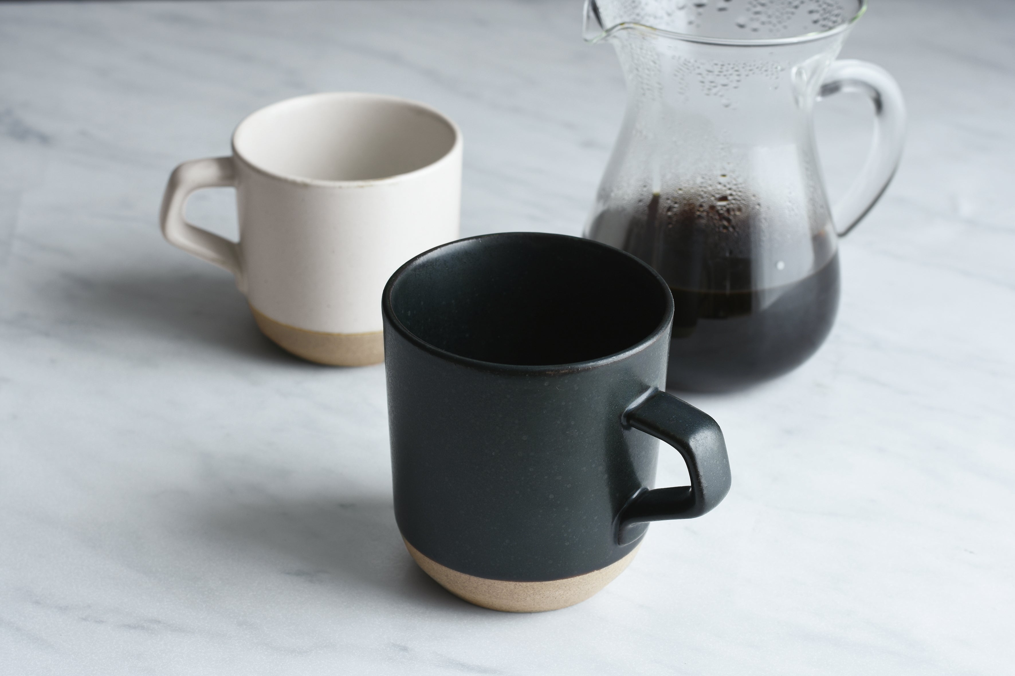 Two ceramic mugs with a glass pitcher of dark coffee on a marble surface.