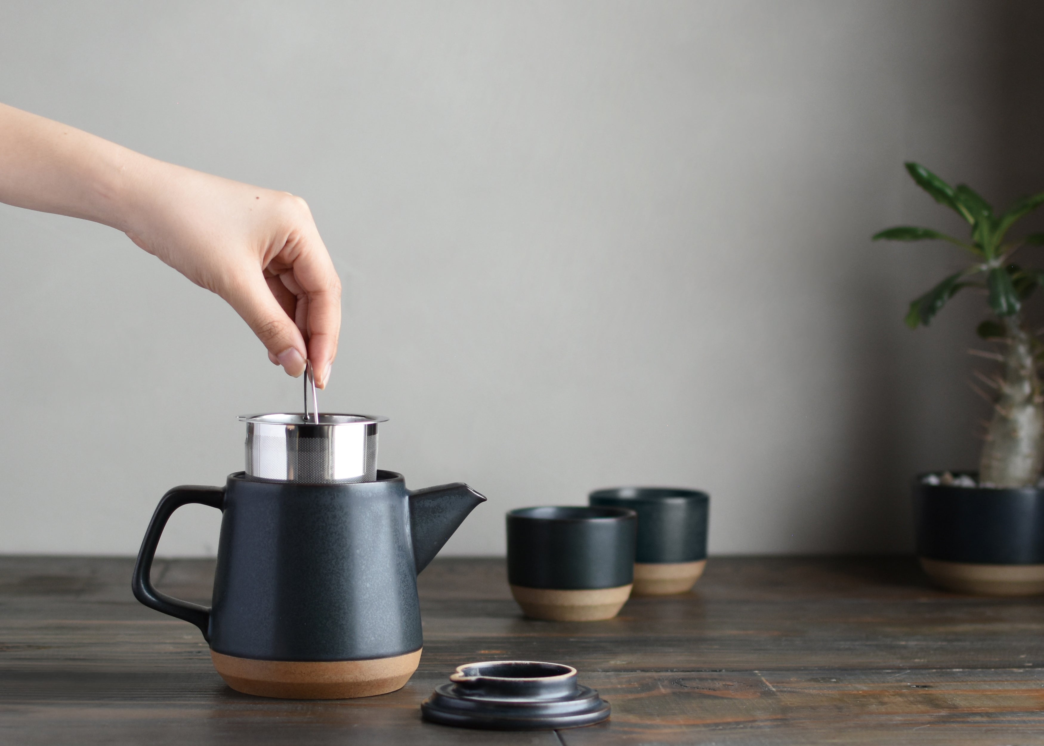 Hand placing a metal infuser into a black teapot on a wooden table, with cups and a plant nearby.