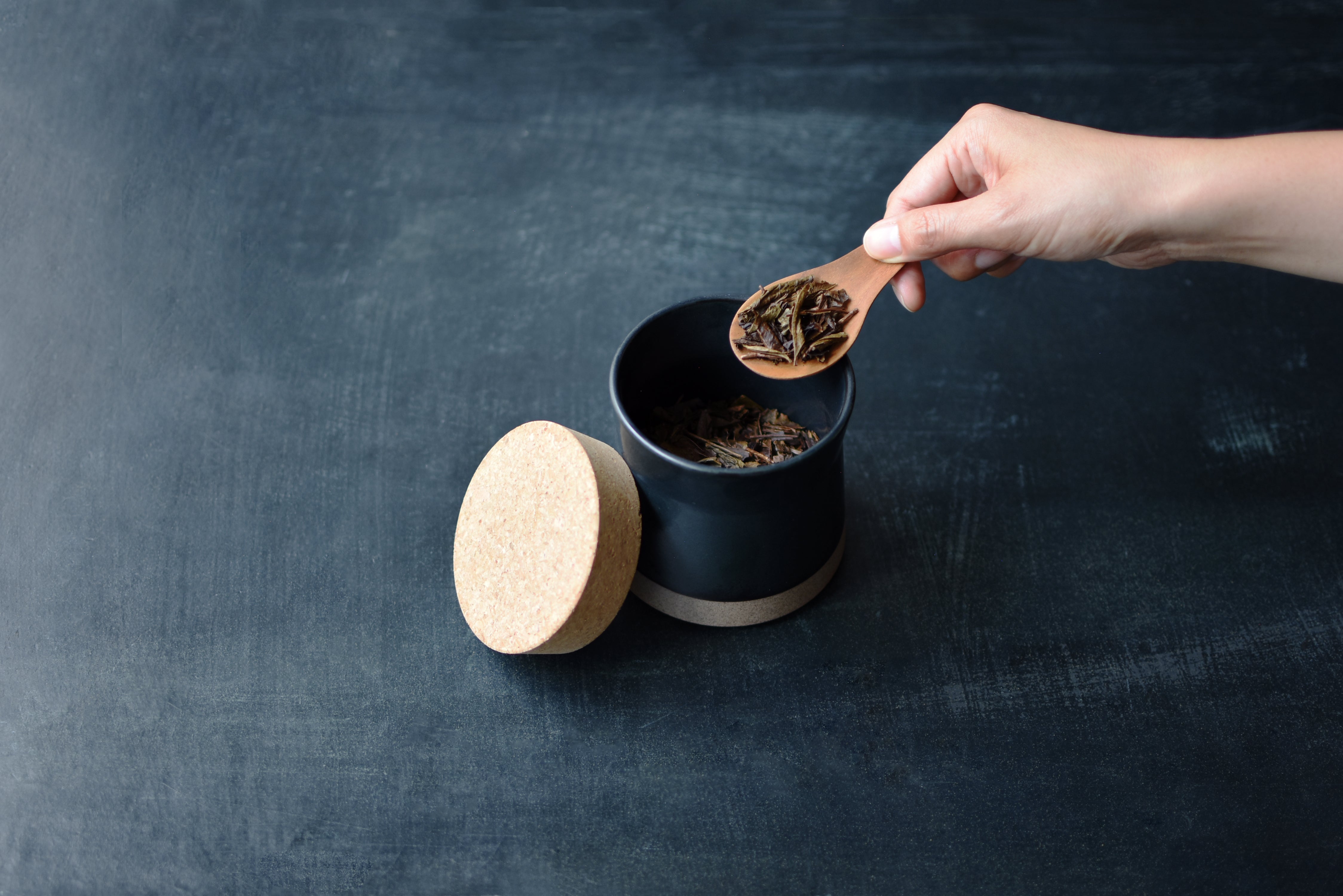 Hand scooping dried herbs from a black ceramic jar with cork lid.