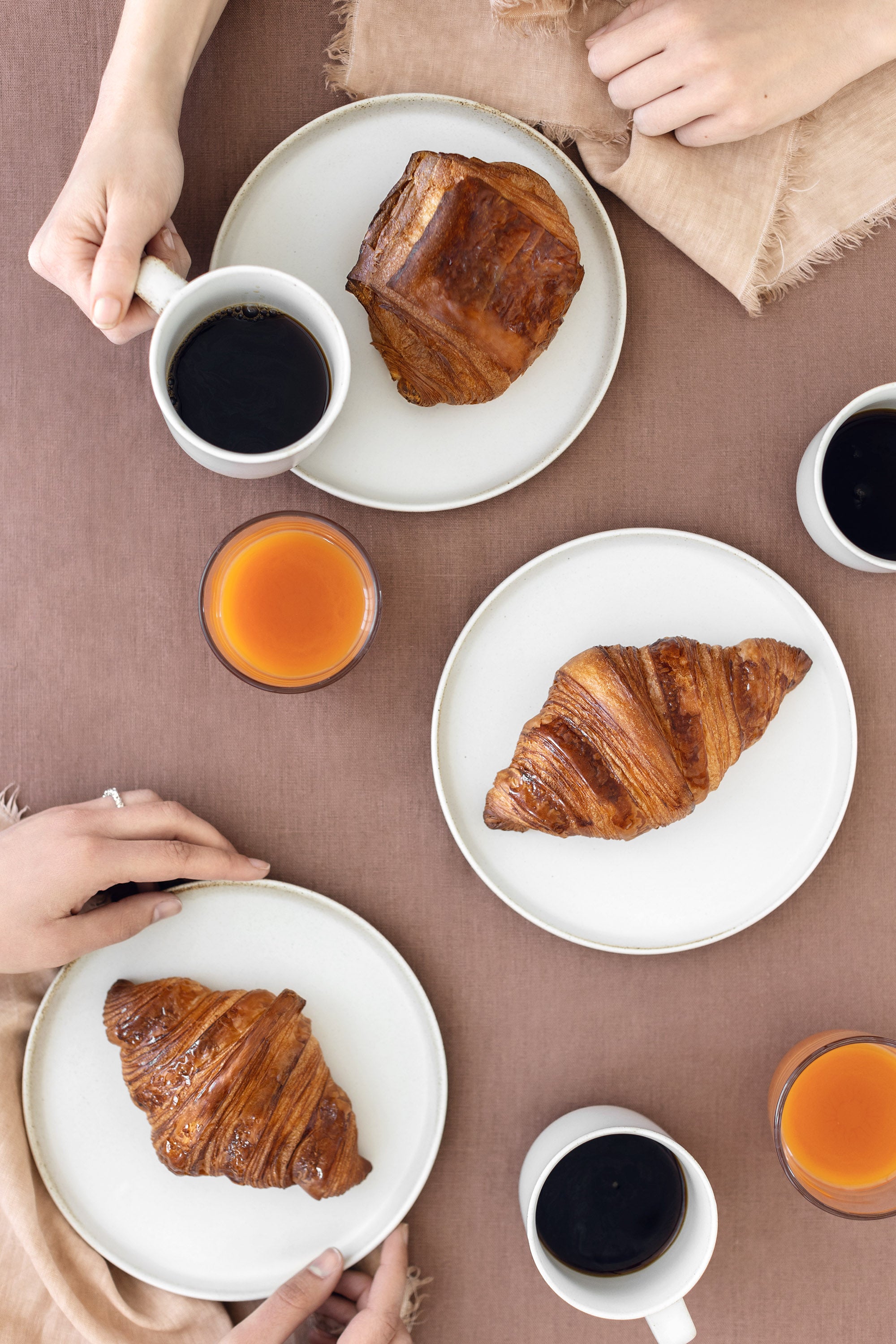 Three croissants and cups of coffee and juice on a table with two hands holding mugs.
