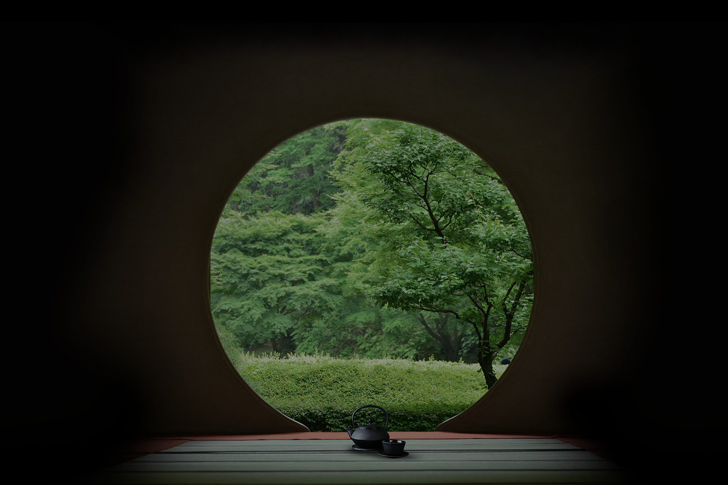 Circular window view of green trees and shrubs with a black teapot on a tatami mat.