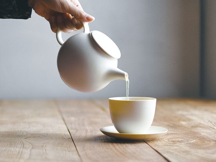 White teapot pouring tea into a cup on a wooden surface with a window in the background.