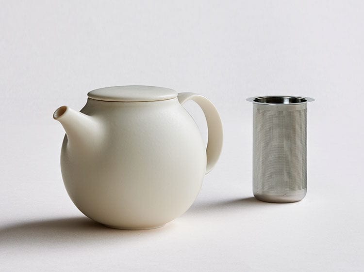 White teapot with lid next to a metal infuser on a plain white background.