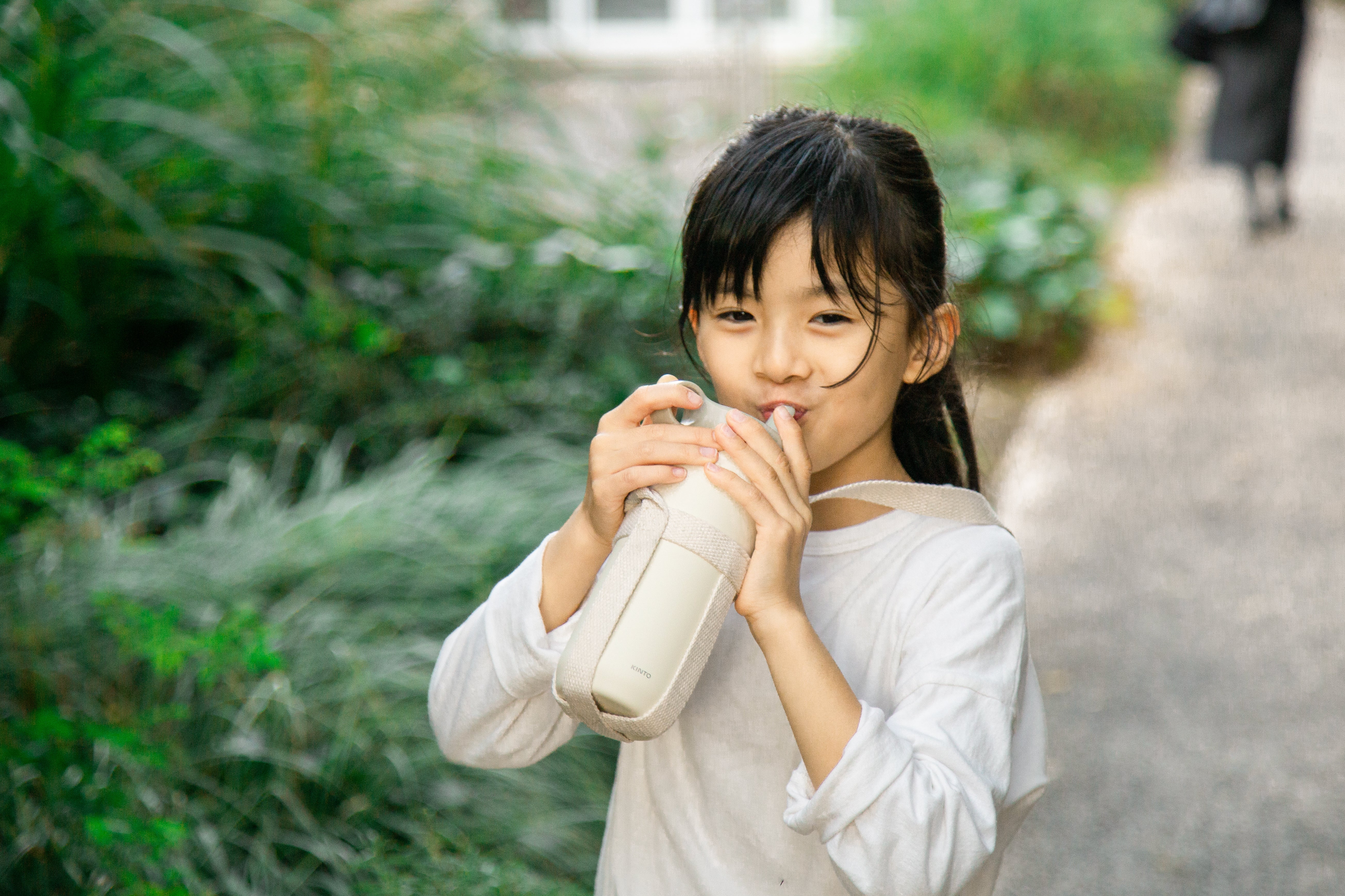 Child in white shirt drinking from a white bottle outside on a path.
