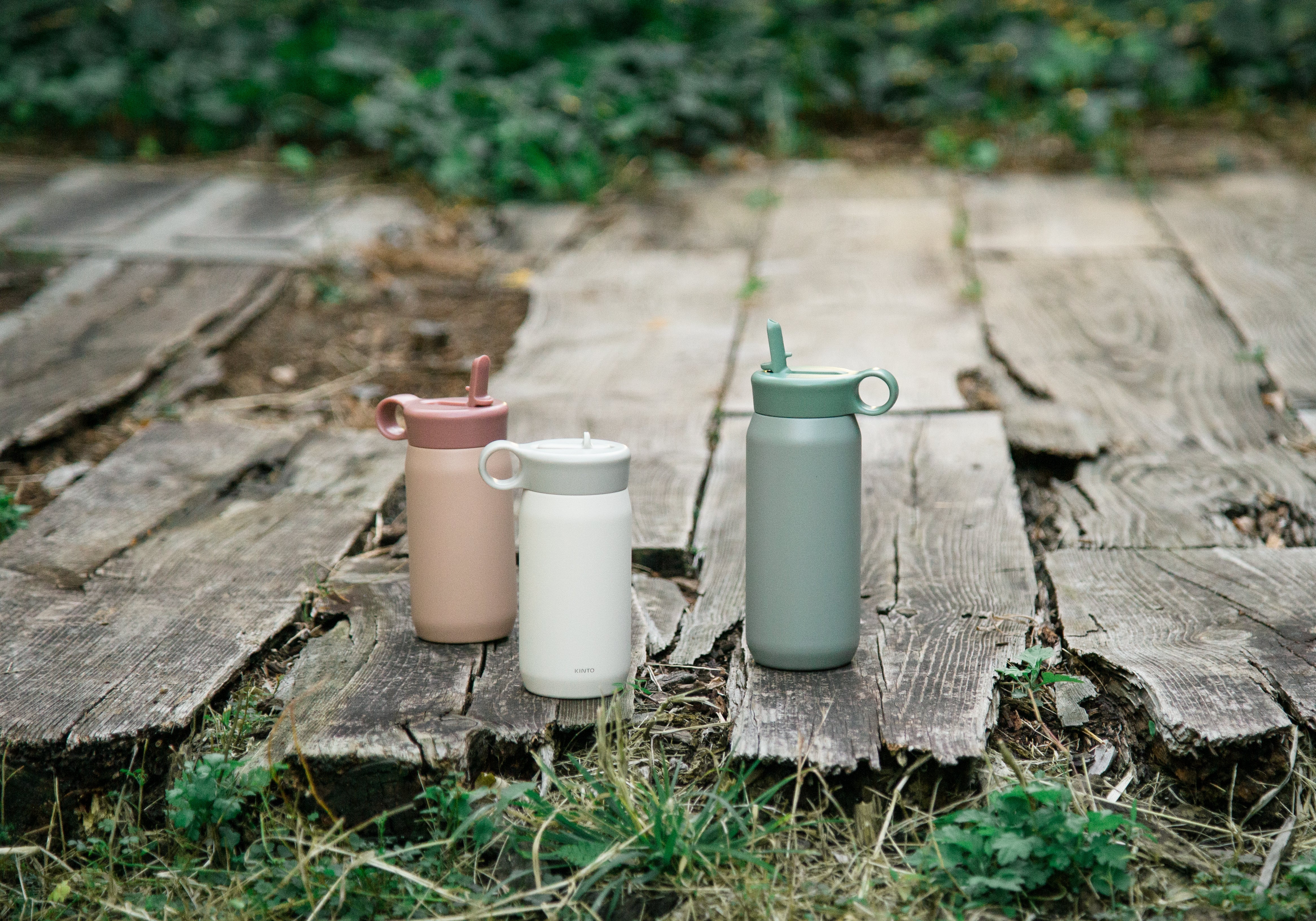 Three bottles in pink, white, and green on wooden planks with grass.