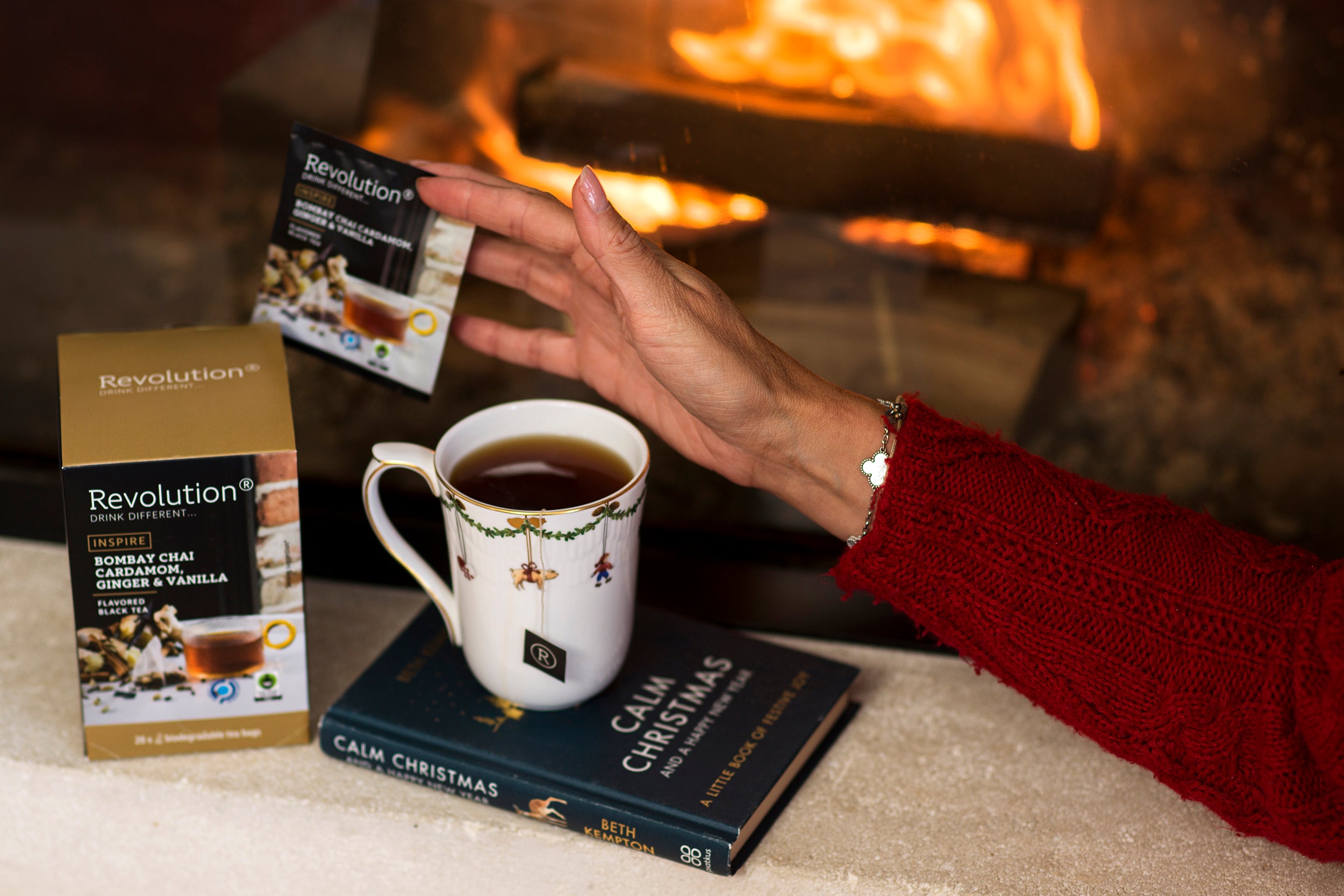 Hand in red sleeve holds tea bag over cup on top of Christmas books by fire.