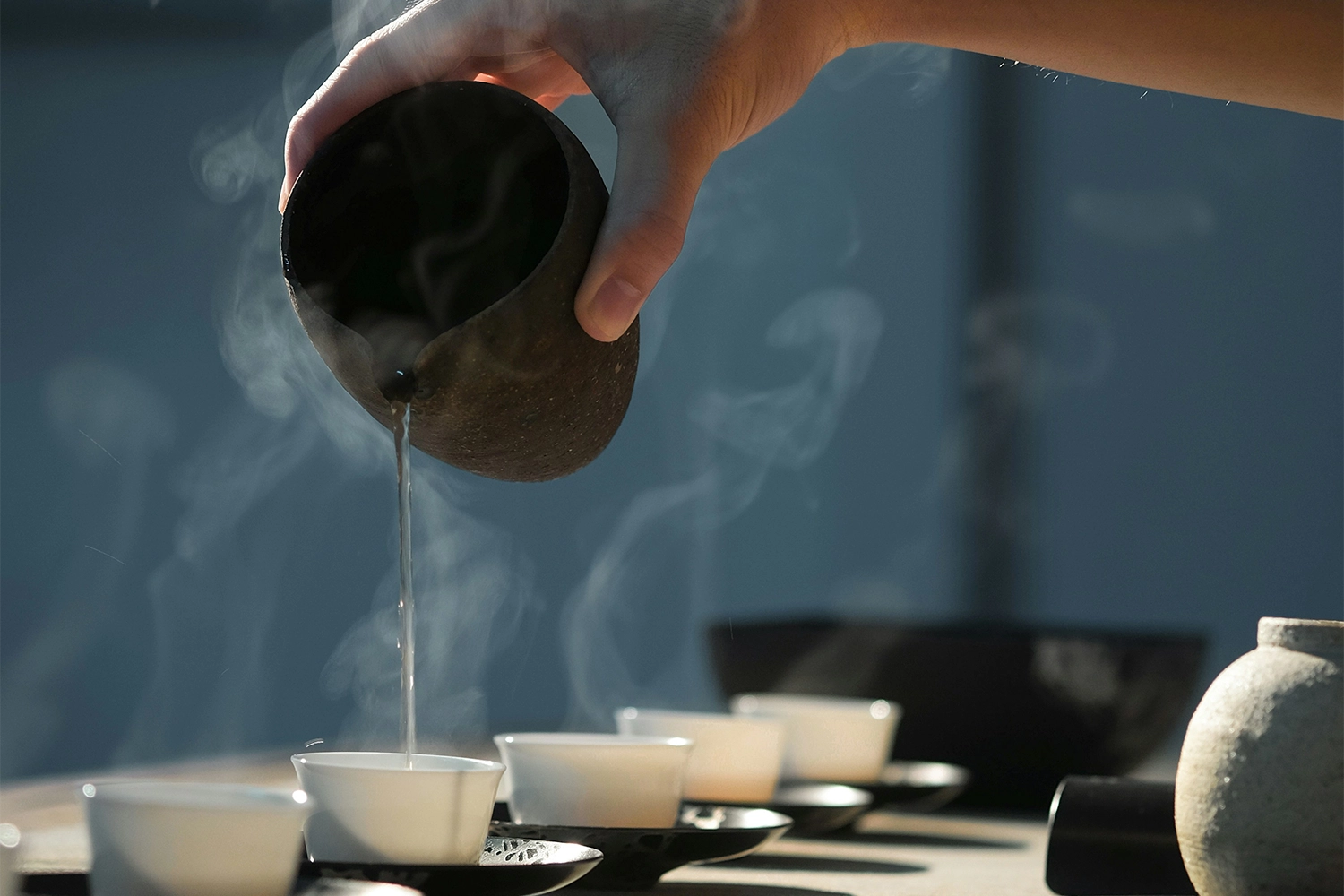 Hand pouring hot water from a dark teapot into small white teacups, with steam rising.