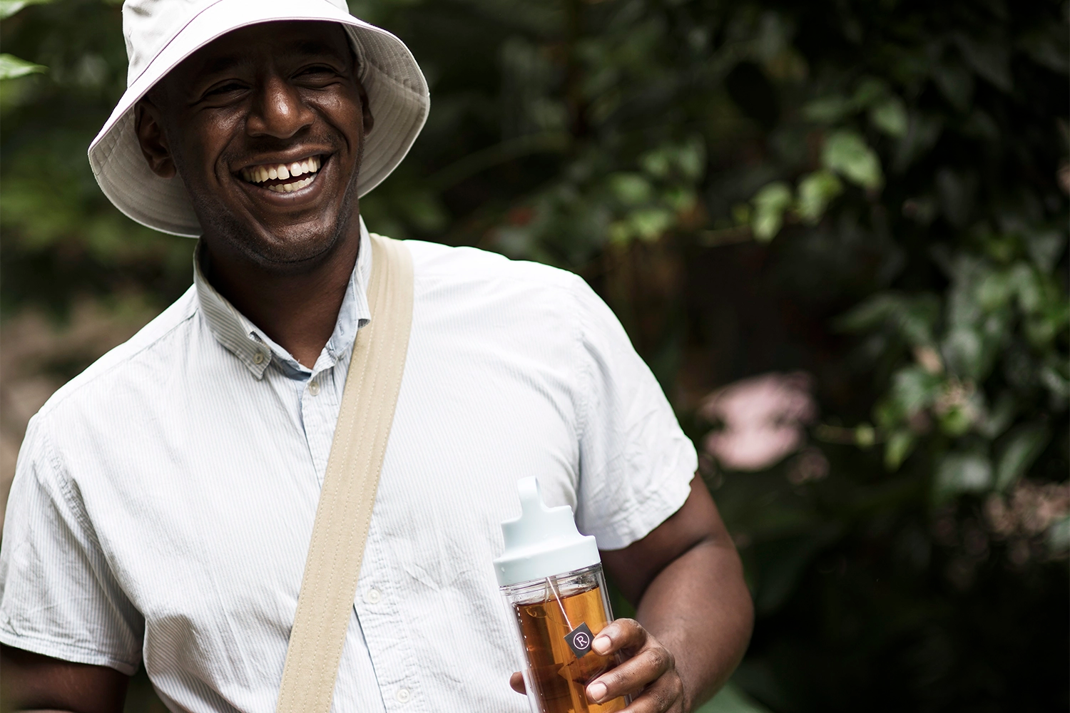 Man in a sun hat and white shirt smiling while holding a bottle outdoors.